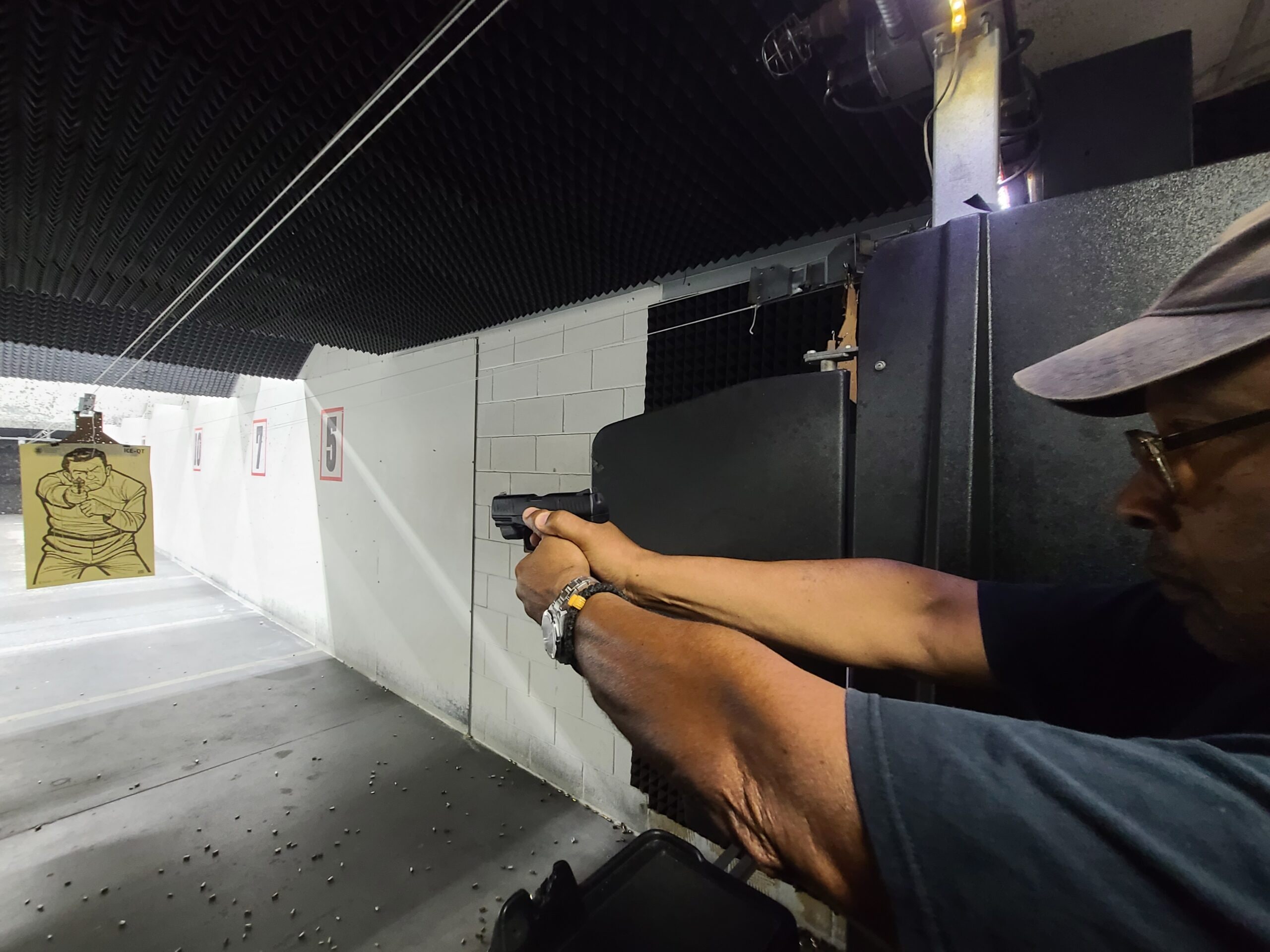 A shooter on the firing line taking aim down an indoor lane, a paper target hanging at the far end