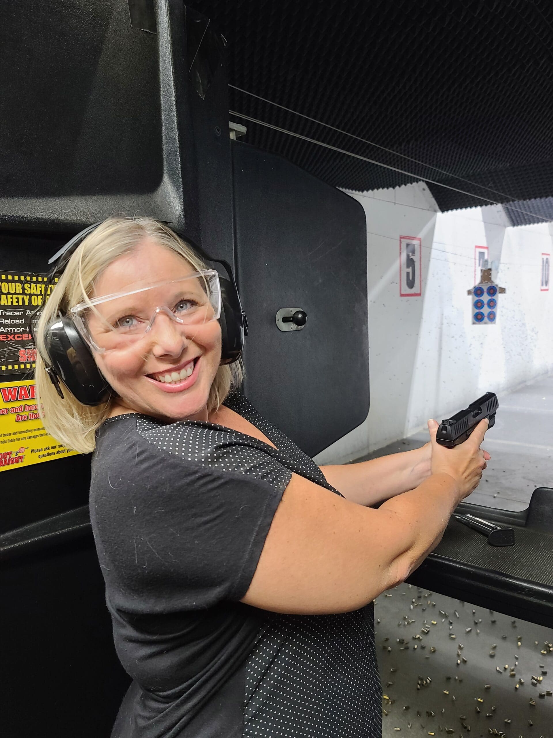 A student in ear defenders and clear safety glasses turns to the camera mid-session, pistol extended toward the five and ten yard markers running away down the lane