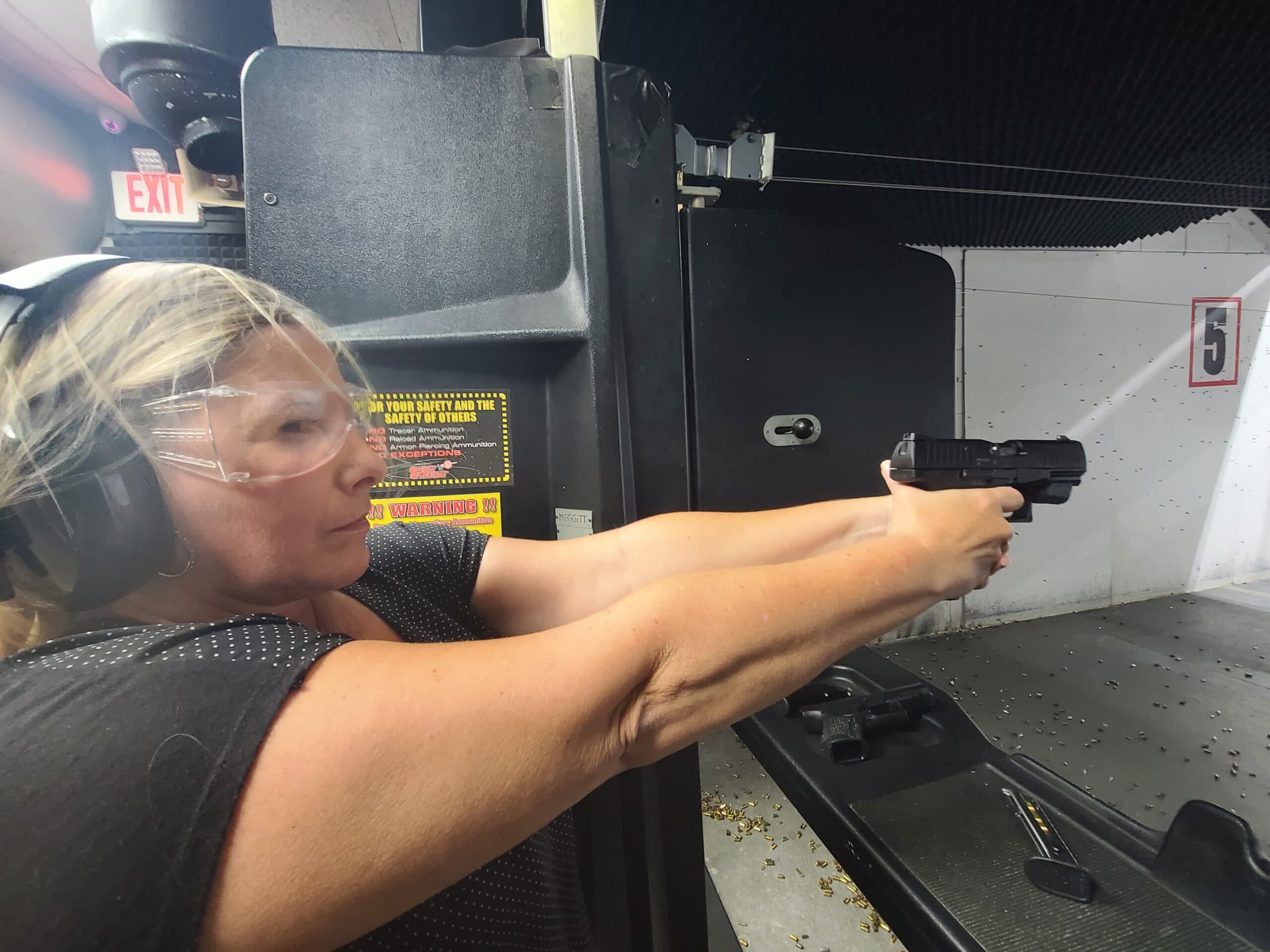 A student in safety glasses and ear defenders holding a pistol at full extension on lane five of an indoor range