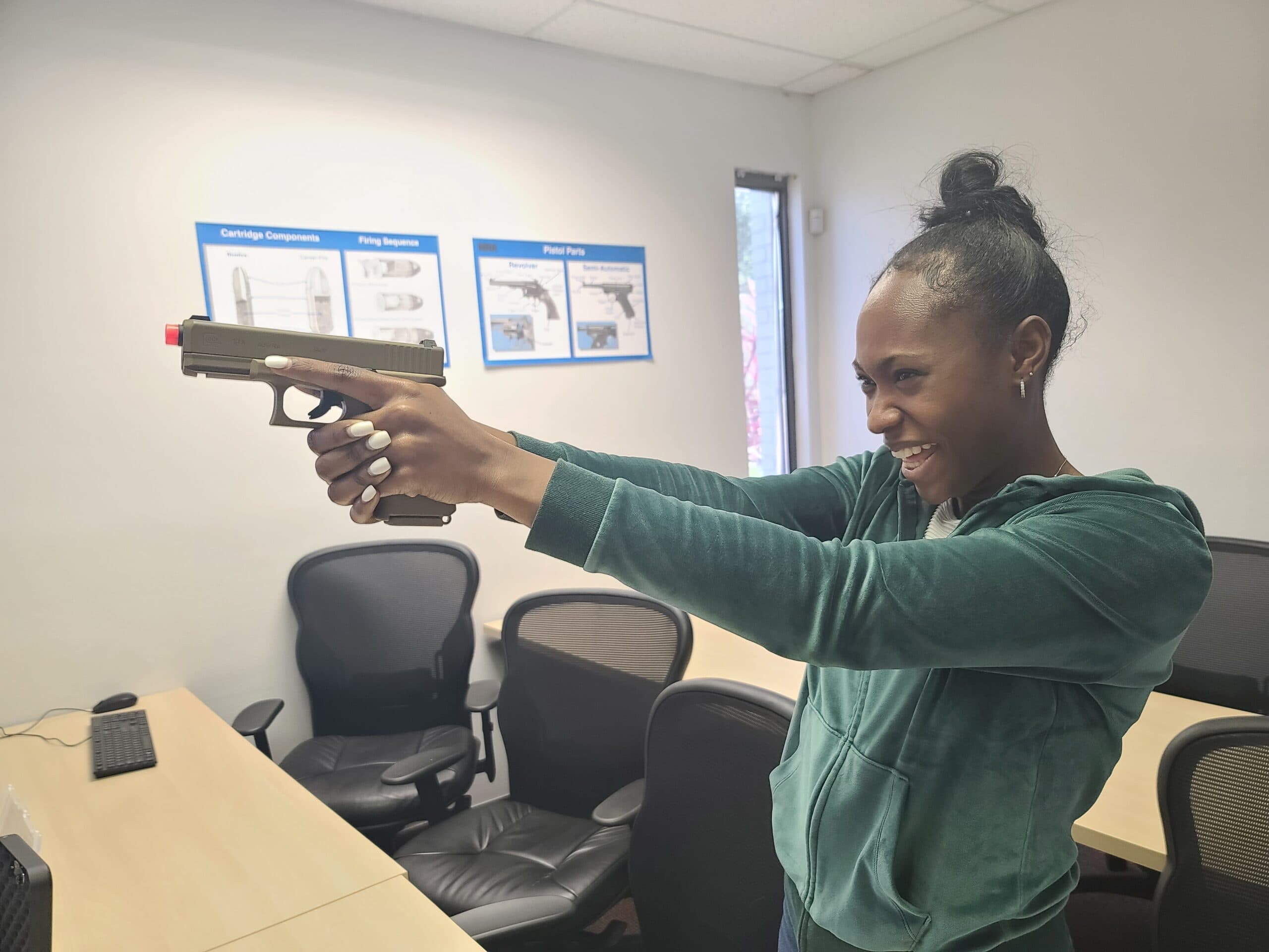 A student practises her stance and grip with an inert training pistol in the National Safety Center classroom, beside the pistol-parts and firing-sequence charts