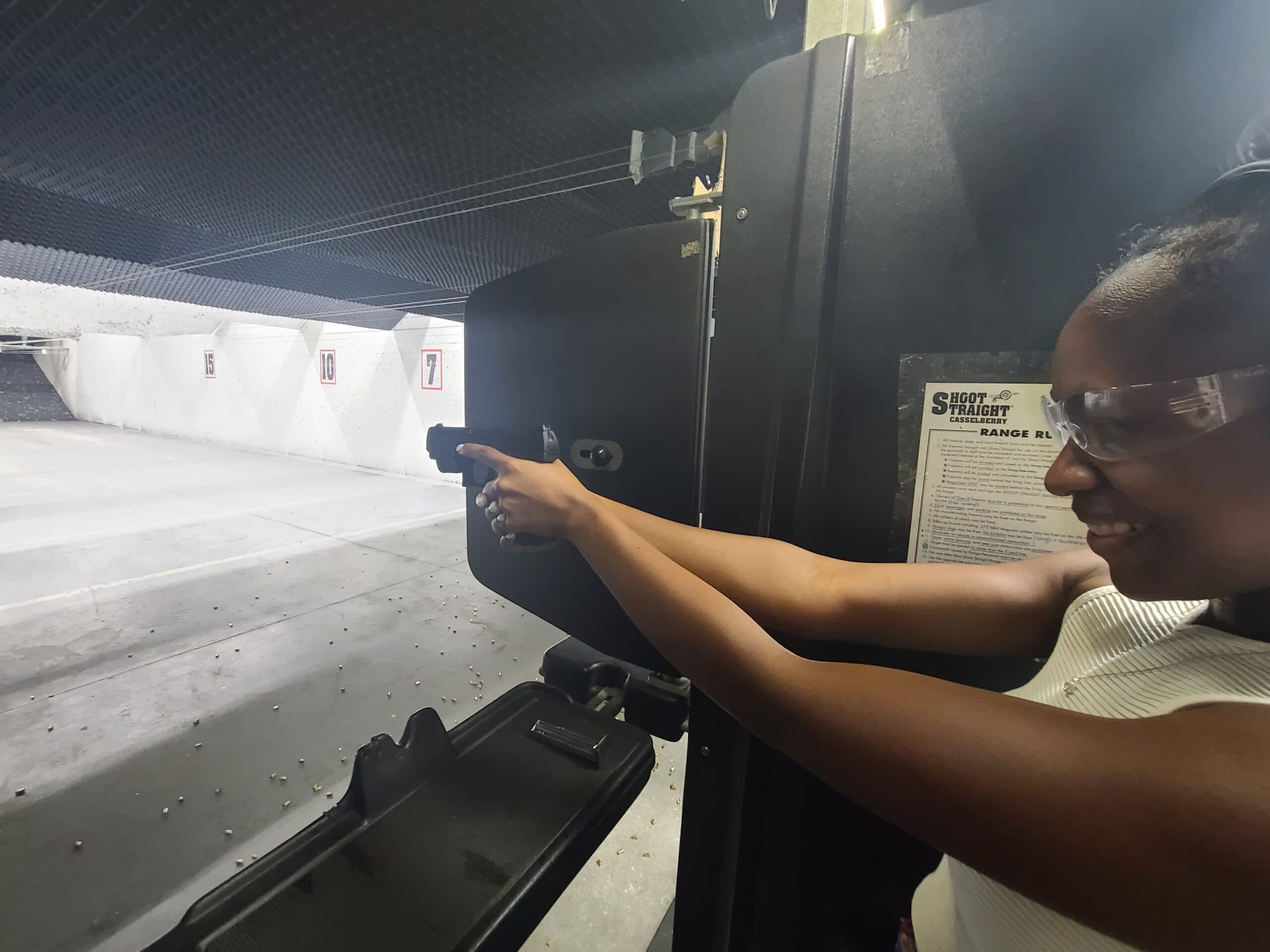 A smiling student in clear safety glasses extends a pistol down a lane at the Shoot Straight range in Casselberry, the seven, ten and fifteen yard markers painted on the wall down the lane