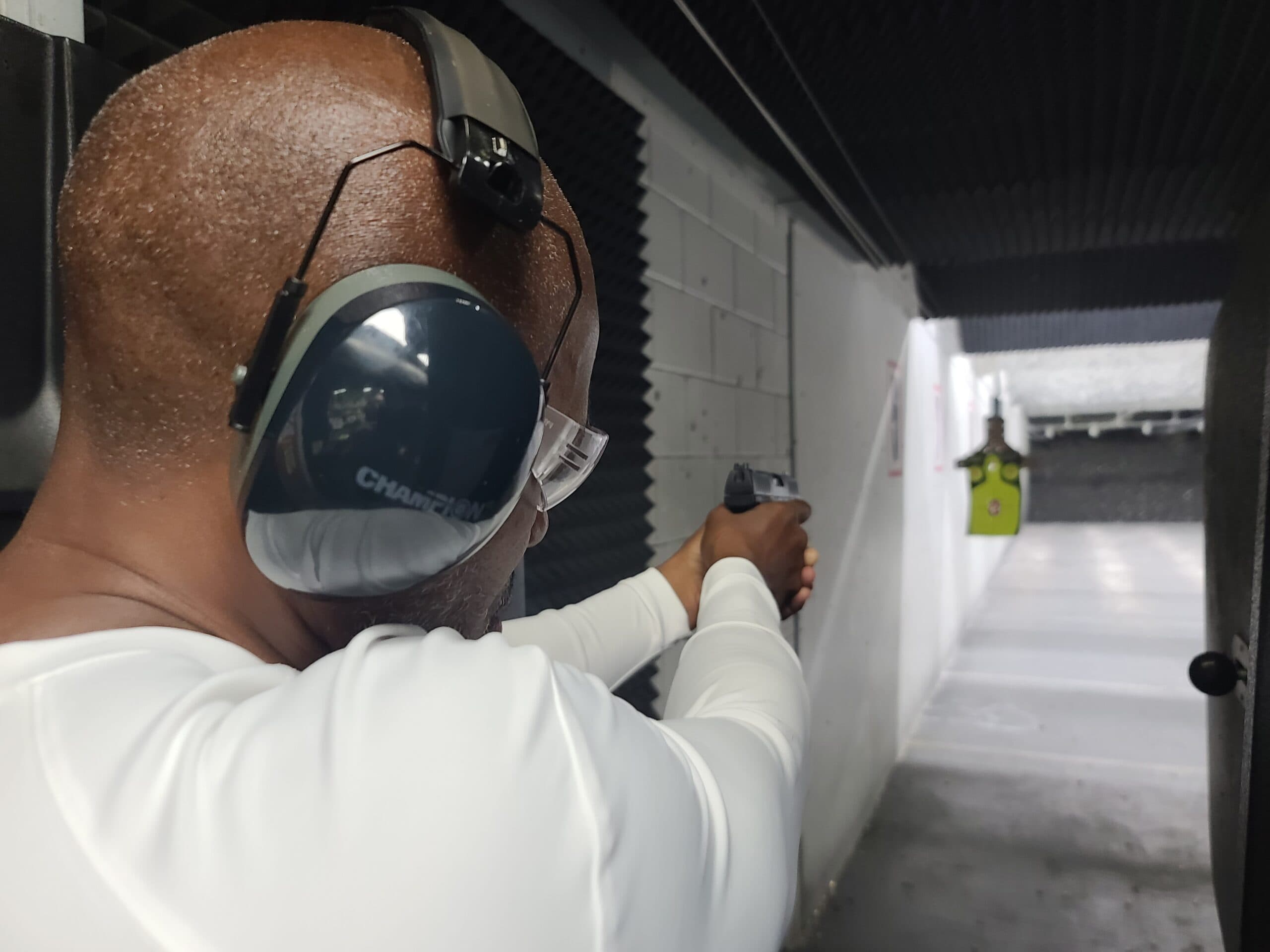 Over a student's shoulder on the firing line, a pistol extended two-handed toward a target hanging at the far end of a brightly lit indoor lane