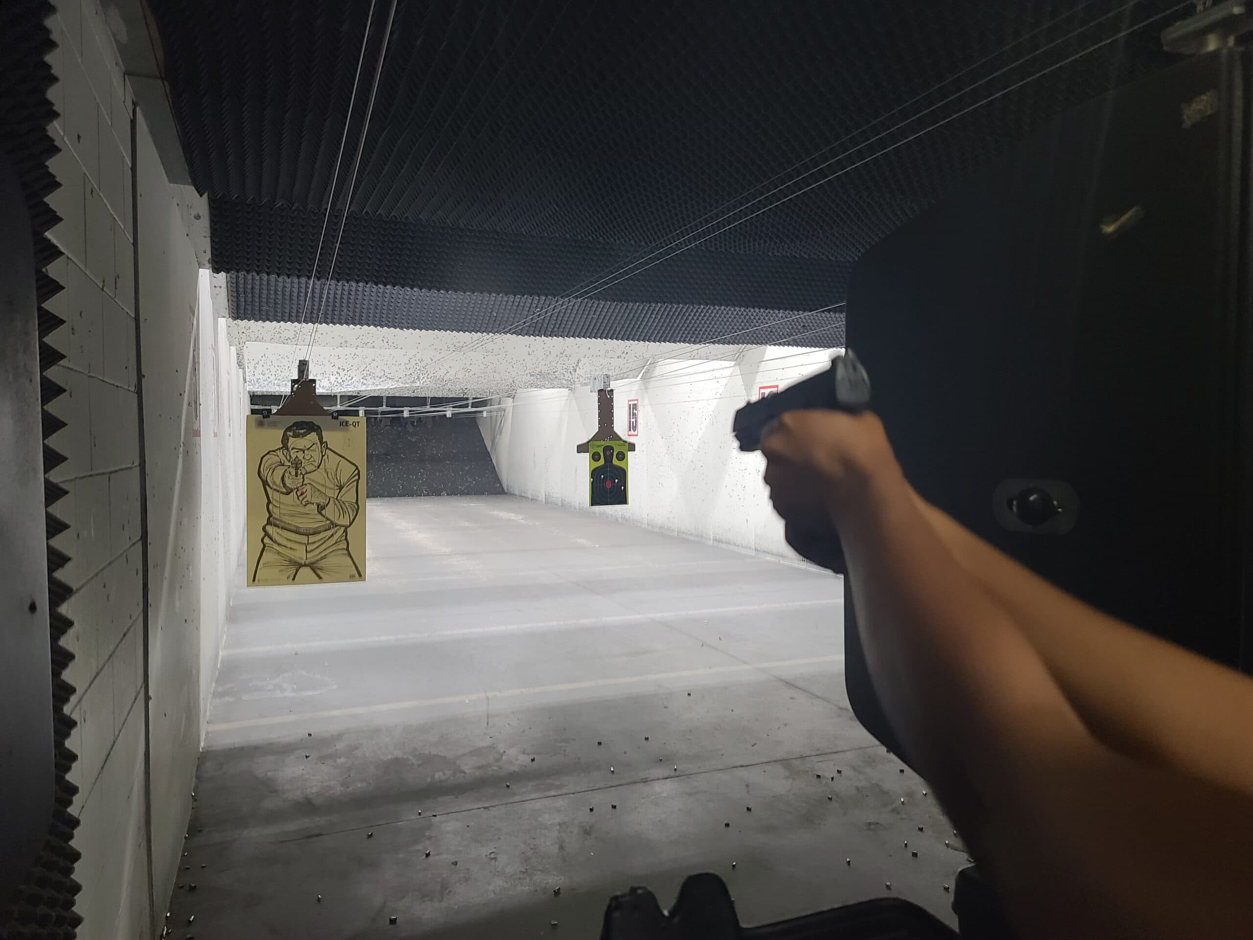 A shooter on the firing line as a spent case flies clear, two targets hanging down the lit lane