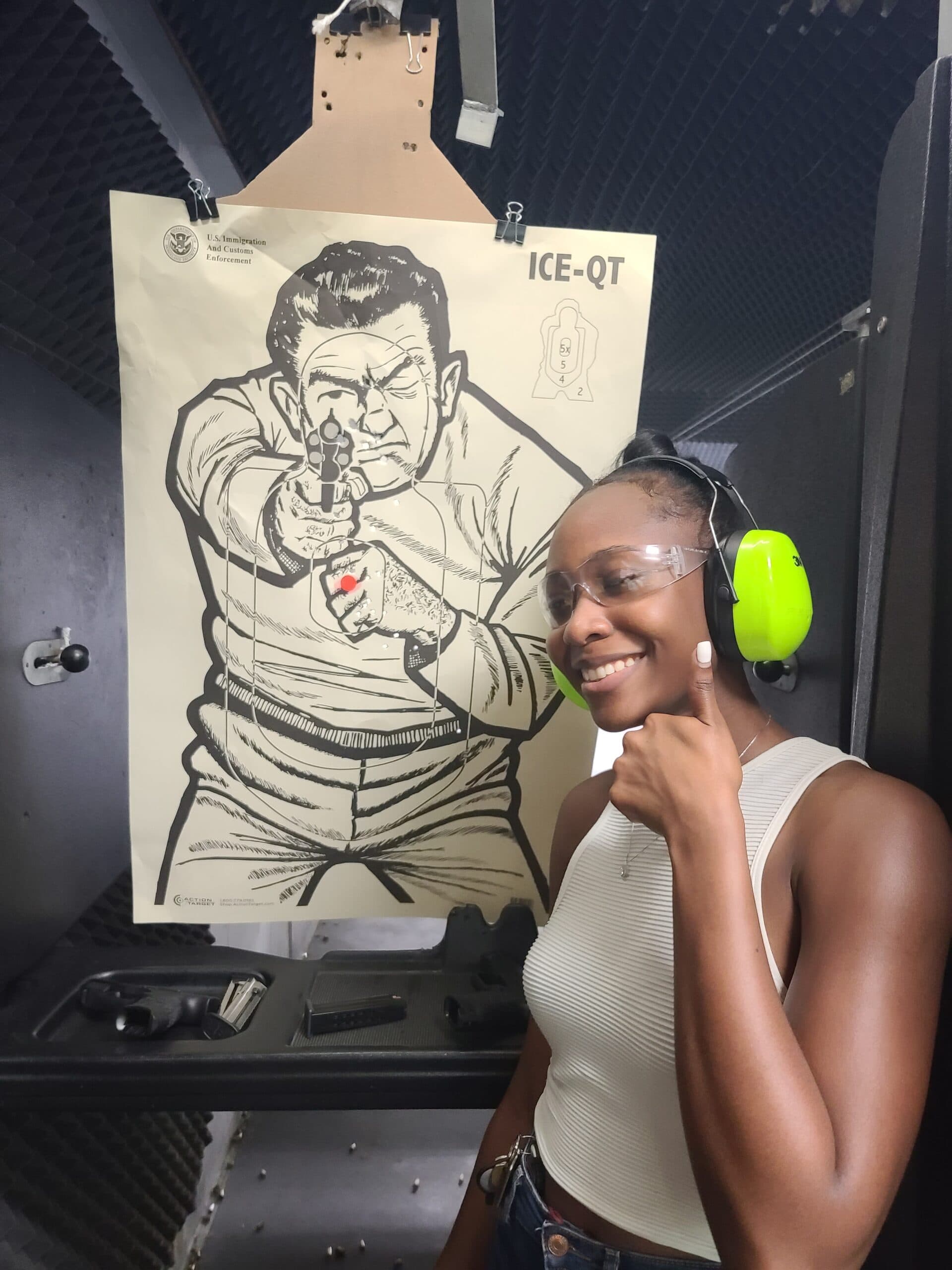 A student in eye protection and bright green ear defenders smiling beside the paper target she has just shot