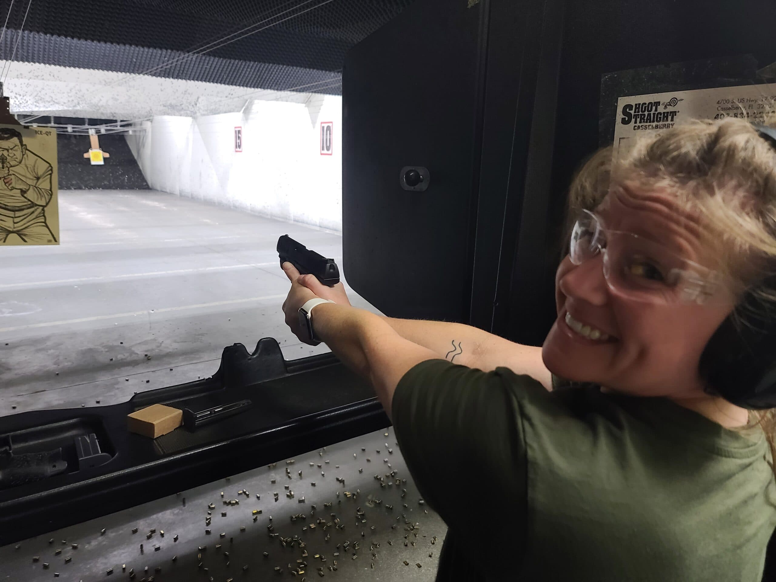 A student in an olive t-shirt looks back over her shoulder, smiling, her pistol still extended down the lit lane at the Shoot Straight range in Casselberry, spent brass covering the booth shelf beside her