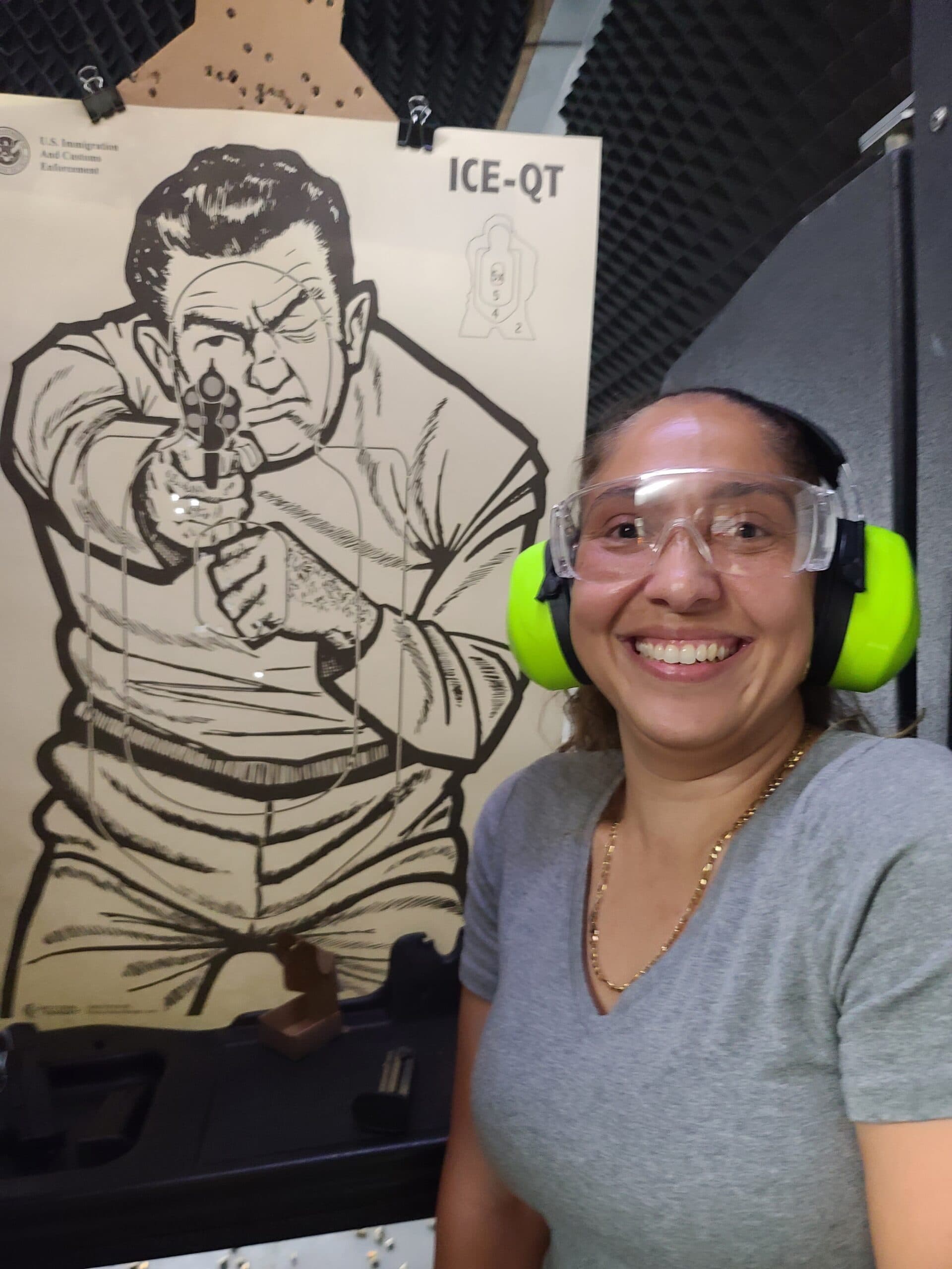 A student in clear safety glasses and green ear defenders smiles beside the target she has just finished shooting