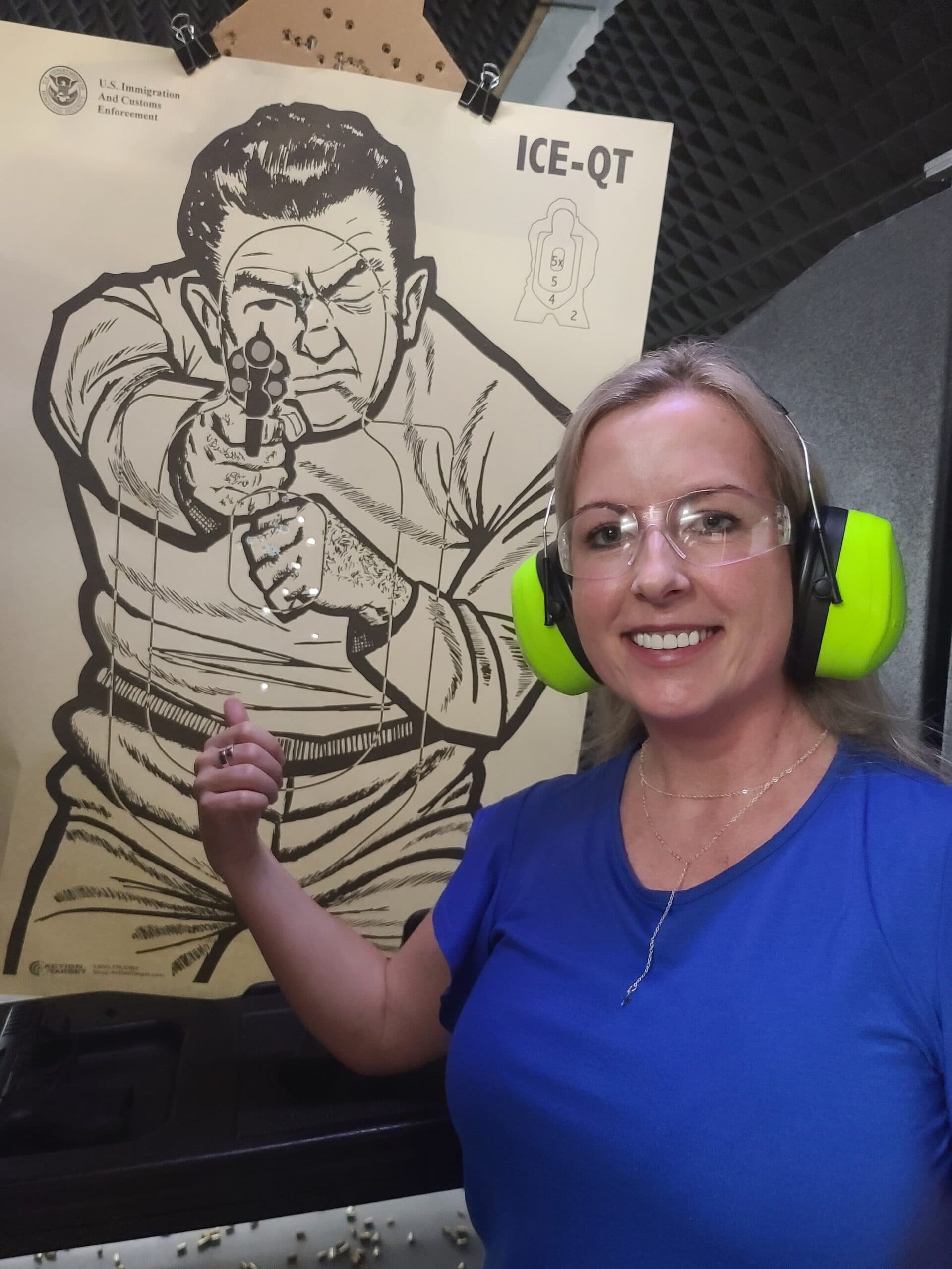 A student in safety glasses and ear defenders gives a thumbs up beside the paper target she has just shot on the range