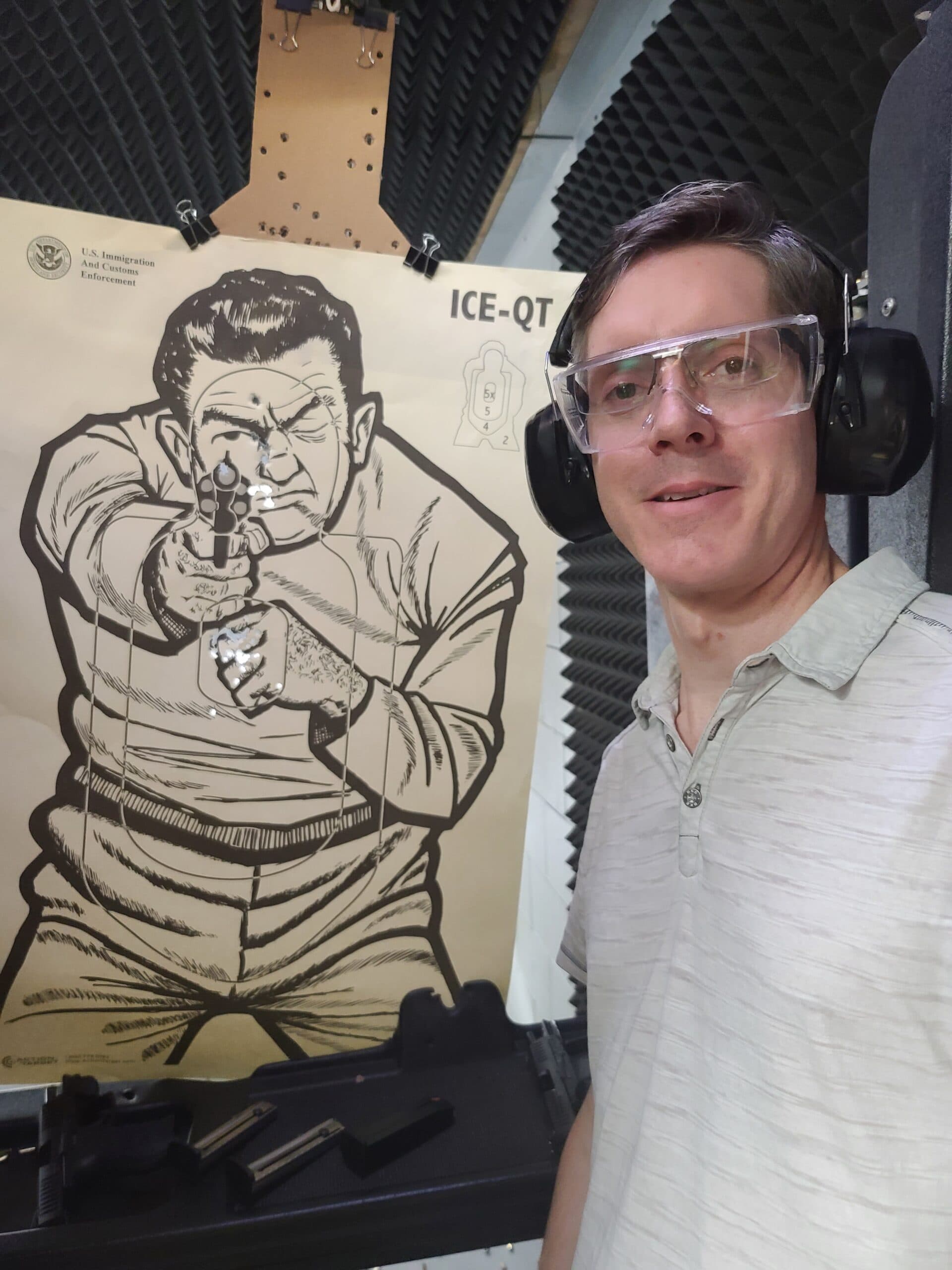 A student in clear safety glasses and ear defenders stands beside the target he has just shot, his pistol laid out on the bench
