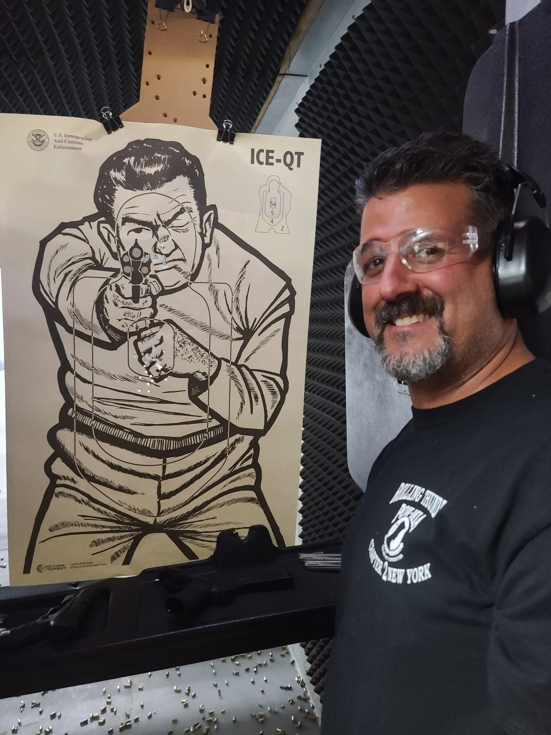 A student grinning next to his target at the end of a range session, wearing safety glasses and ear defenders
