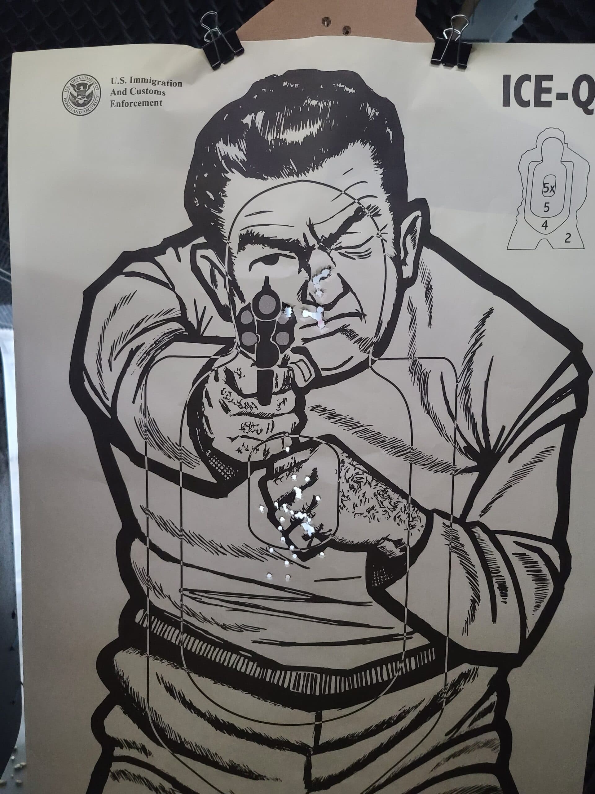 A shot-up ICE-QT paper target still clipped to its carrier, one tight group punched through the chest and a scatter of holes across the face