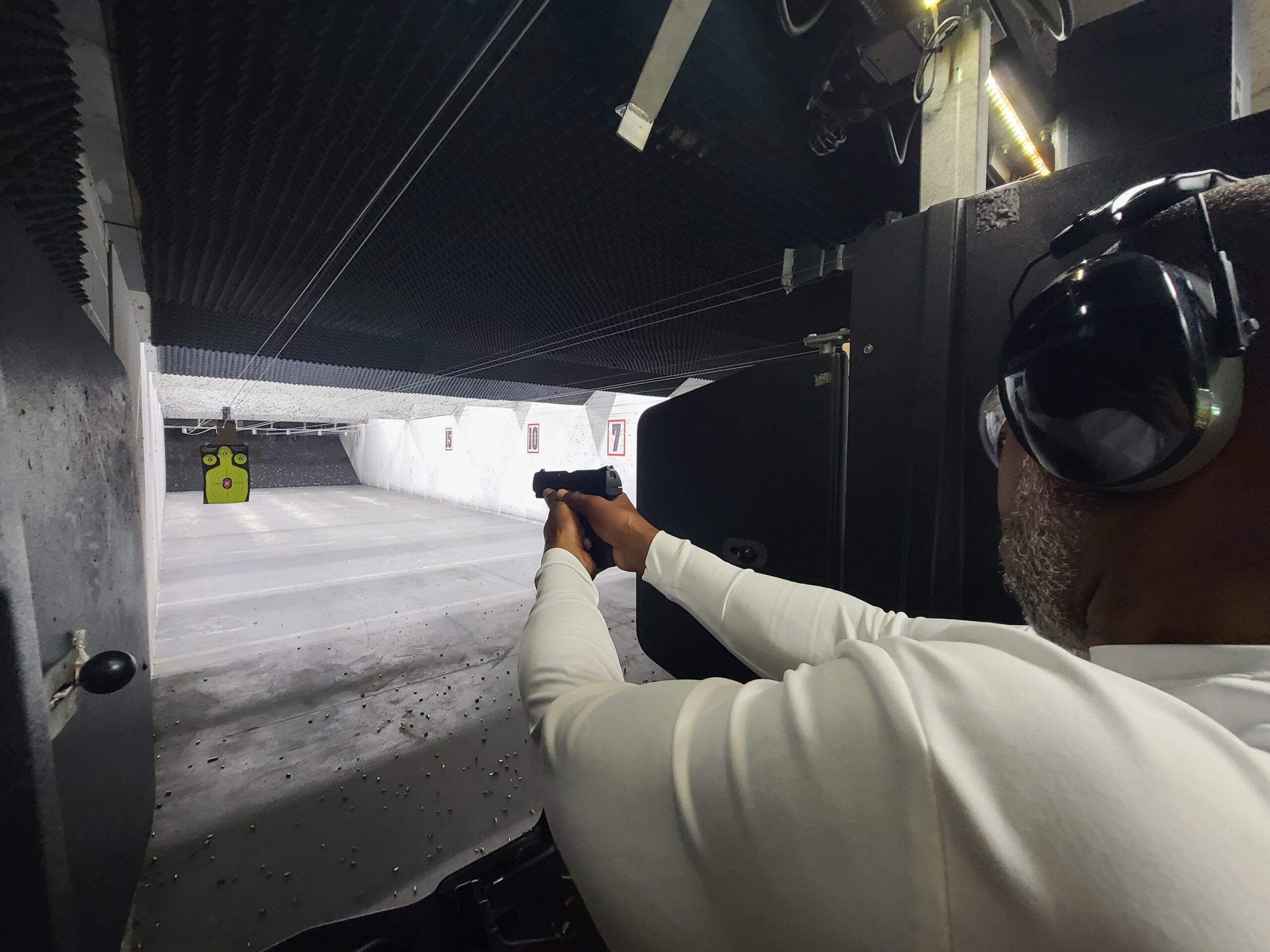 A student in eye and ear protection holds a two-handed grip on the firing line, a target hanging at the far end of the lit lane