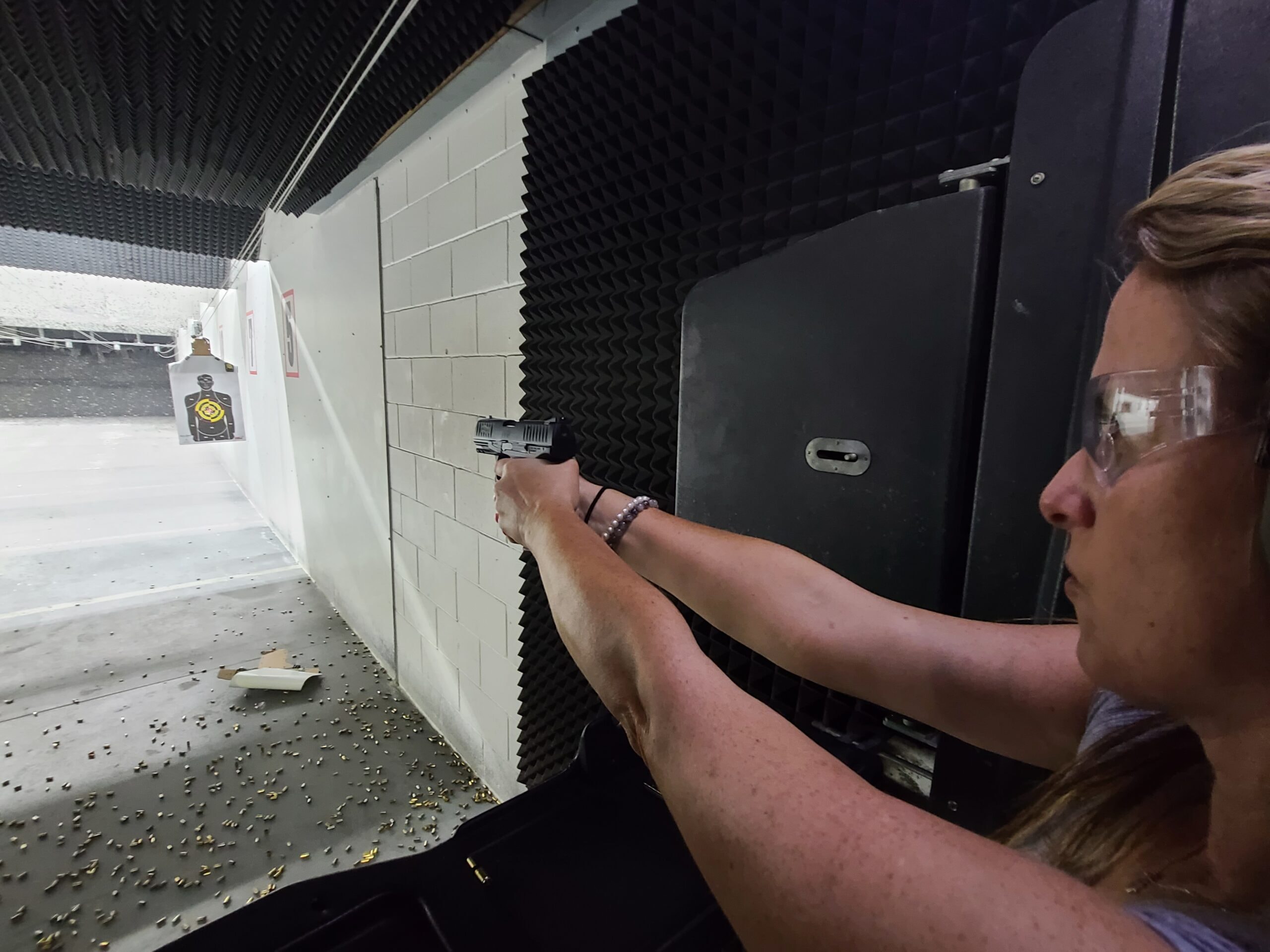 A student firing a pistol from an indoor booth, the lit lane running away downrange to a paper silhouette target, spent brass scattered across the floor